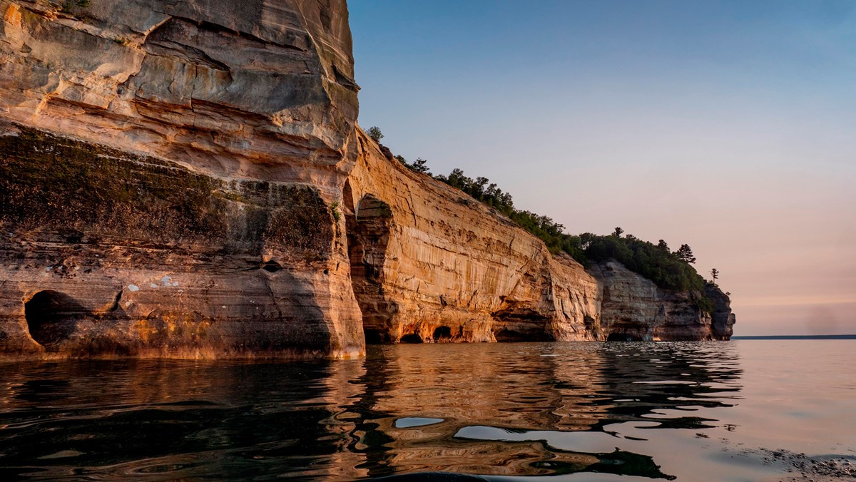 Sandstone cliffs with streaks of brown, red, and orange rise out of Lake Superior at sunset. 