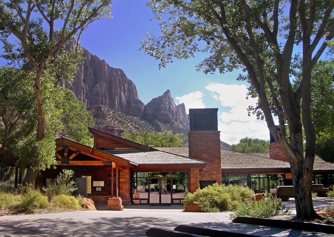 A sandstone and glass building surrounded by tall trees with a mountain behind it. 