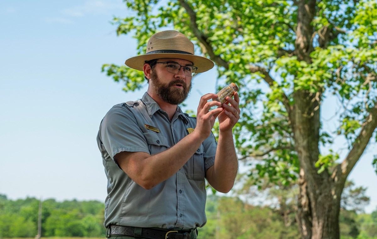A park ranger holds up a specimen of limestone as he speaks outdoors.