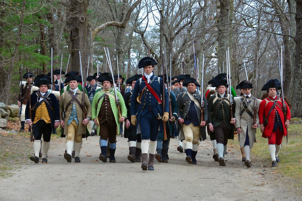 A group of militia soldiers standing shoulder to shoulder with muskets march down a dirt road.