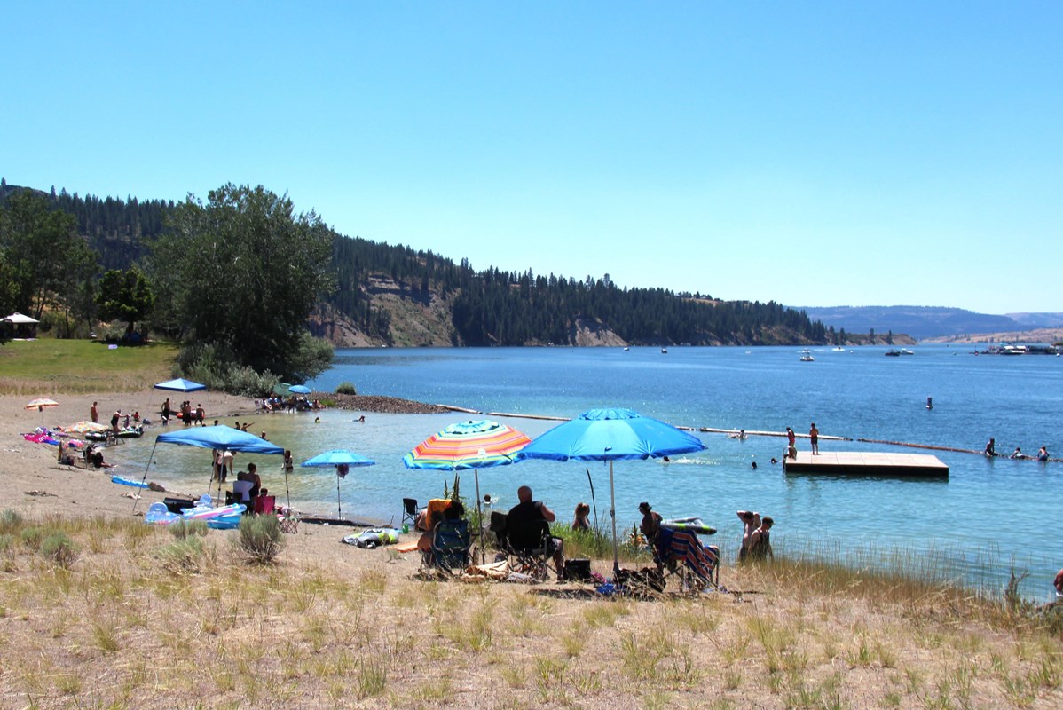 Scattered beach umbrellas shade various people sitting in chairs at a lakeside beach.