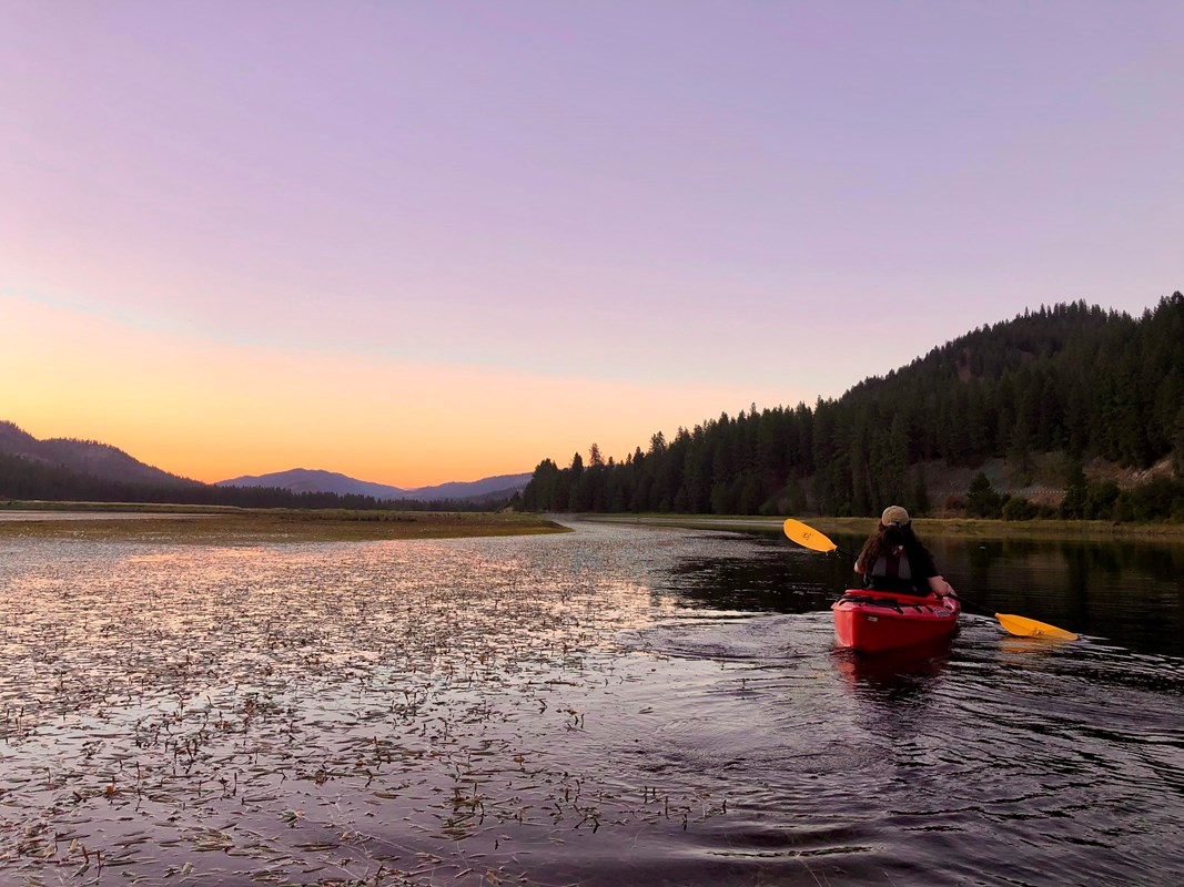 Warm and purple hues light up the sky and water as a kayaker in a red kayak paddles along.