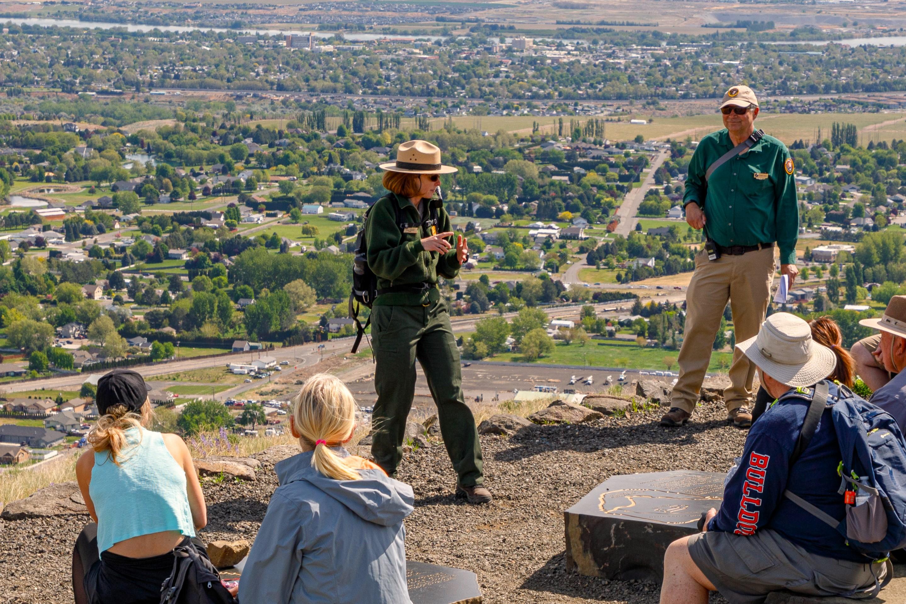 Uniformed park ranger talks animatedly to visitors on a hillside overlooking a sprawling urban area.