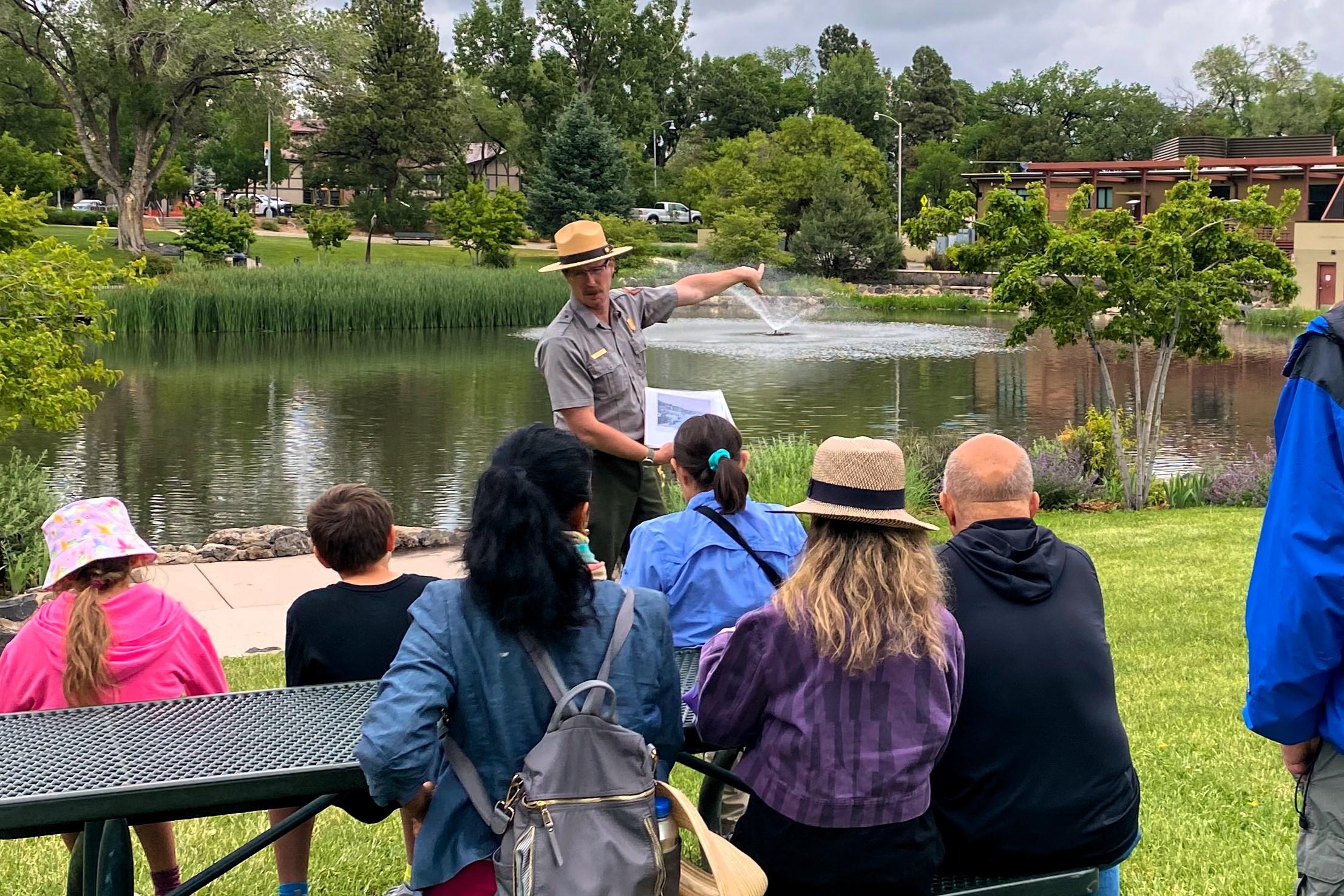 Uniformed park ranger points to distance of fountain and trees as visitors sit enraptured.