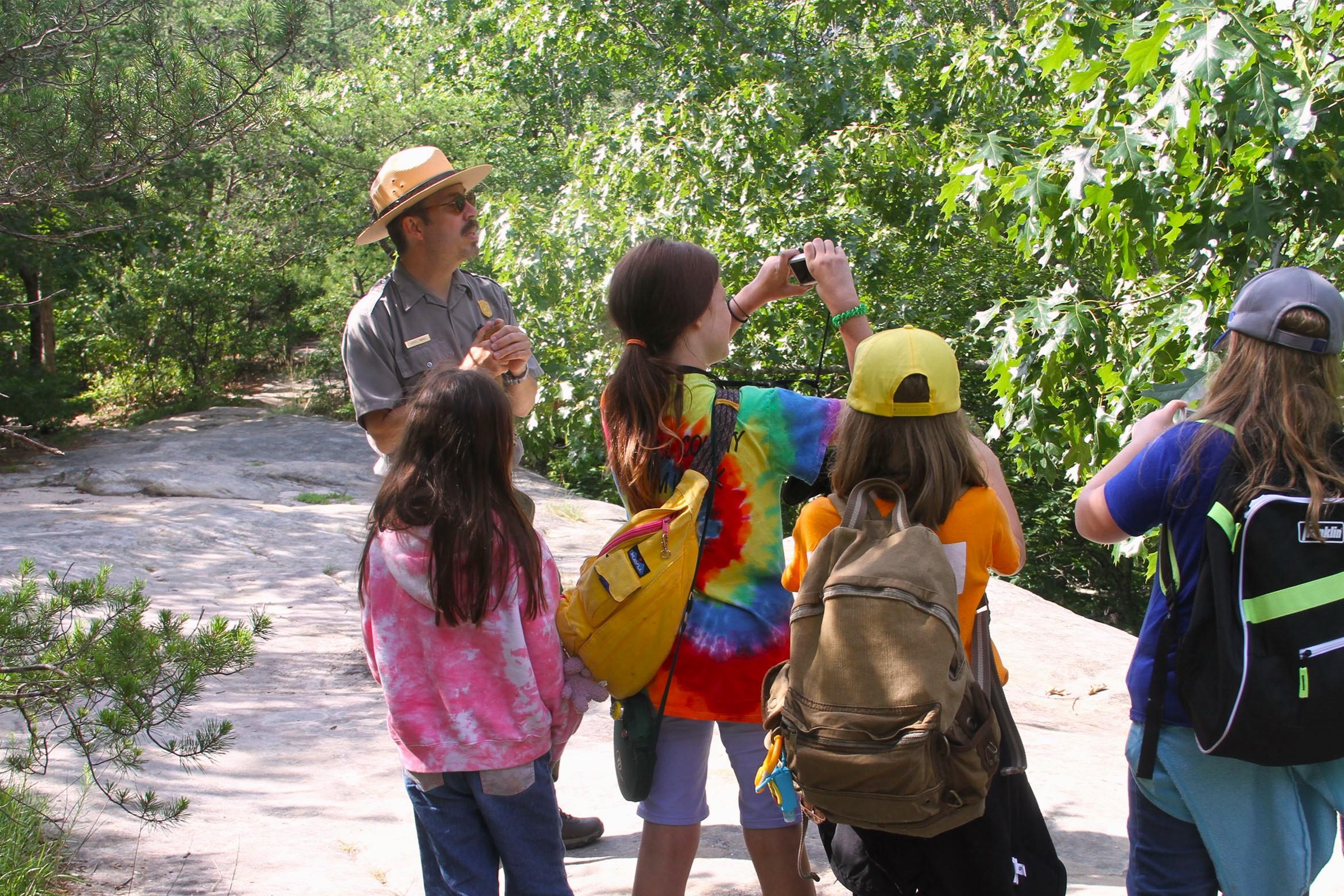 Uniformed park ranger stands with youth along path lined with green broadleaved trees.