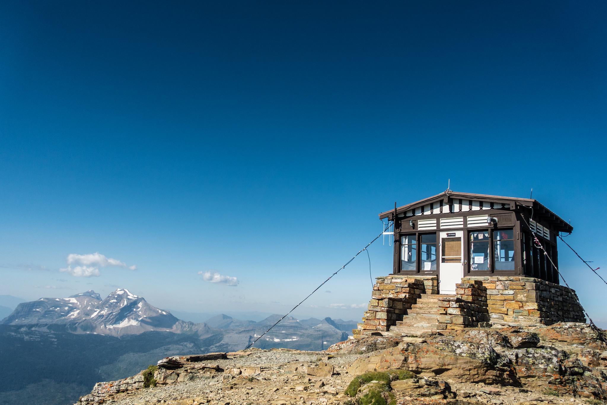 A small, brown and white structure sits on a rocky top with mountain peaks in the distance.