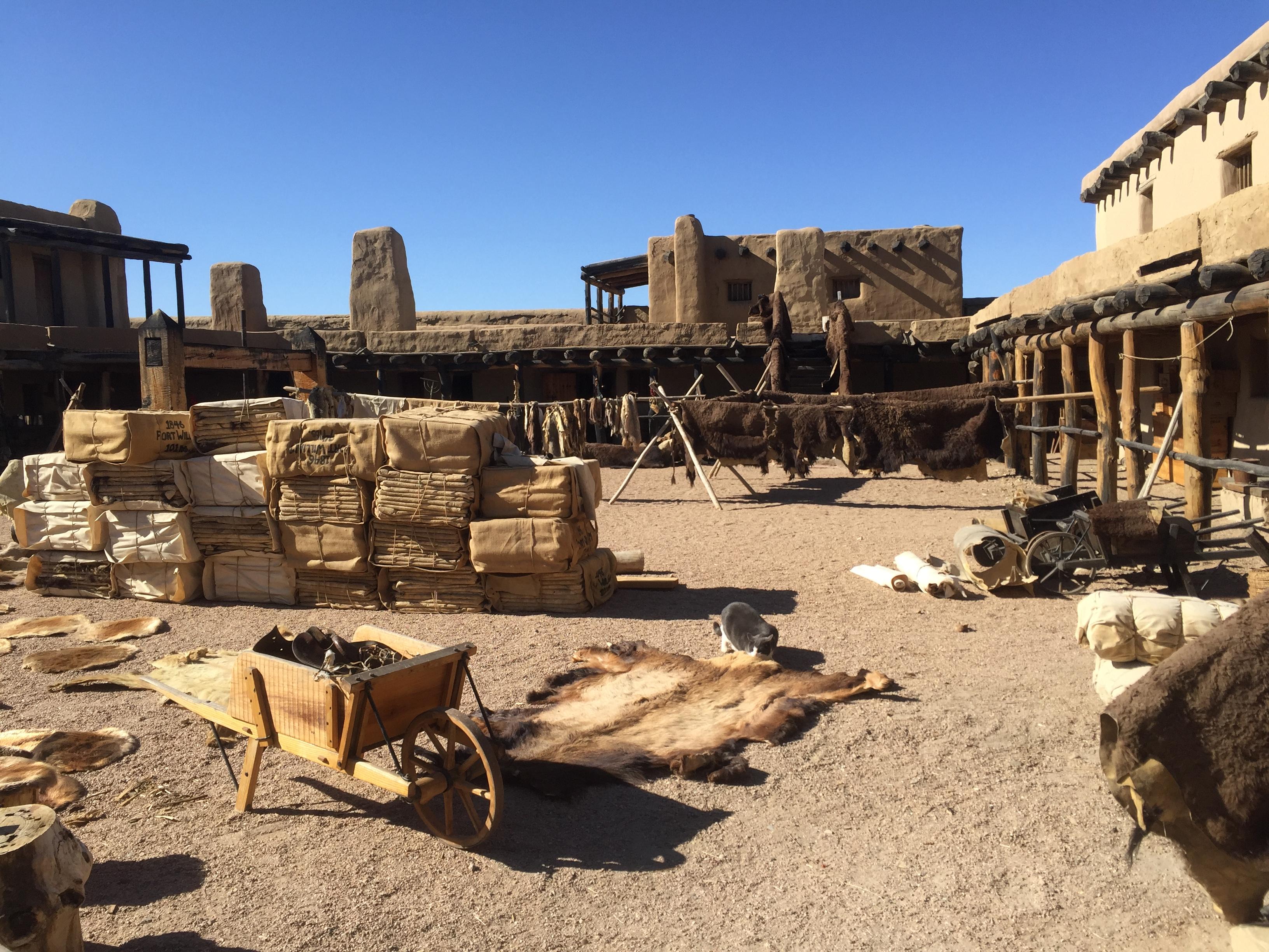 Wooden boxes and buffalo furs are scattered around a plaza inside an adobe fortification.