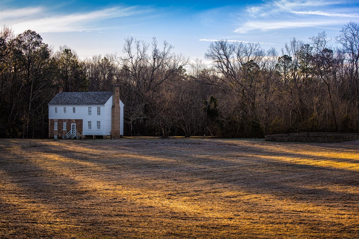 A rectangular house of wood and brick, with a cedar shingle roof and brick chimney, near woods. 