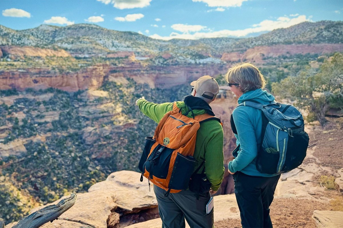 two people stand together at canyon cliff edge pointing to tree covered hills across the canyon