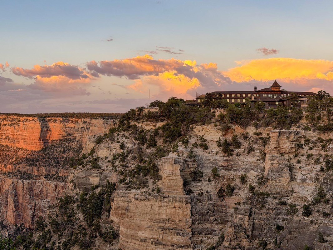 Colorful sunset clouds in the sky above a rustic, four-story hotel on the edge of a precipice.