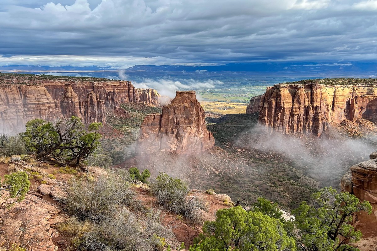 a wide sandstone monolith stands next to cliffs on a misty, cloudy day with green shrubs below