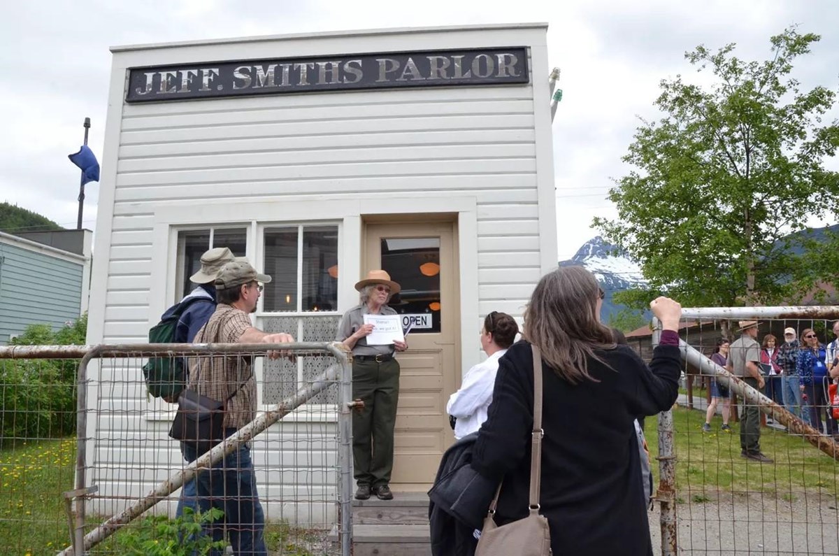 A park ranger and visitors stand in front of a small white building reading "Jeff. Smiths Parlor"