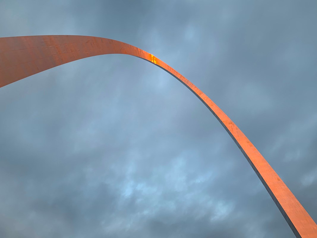 looking up at the Gateway Arch against a cloudy blue sky with a orange reflection at the top