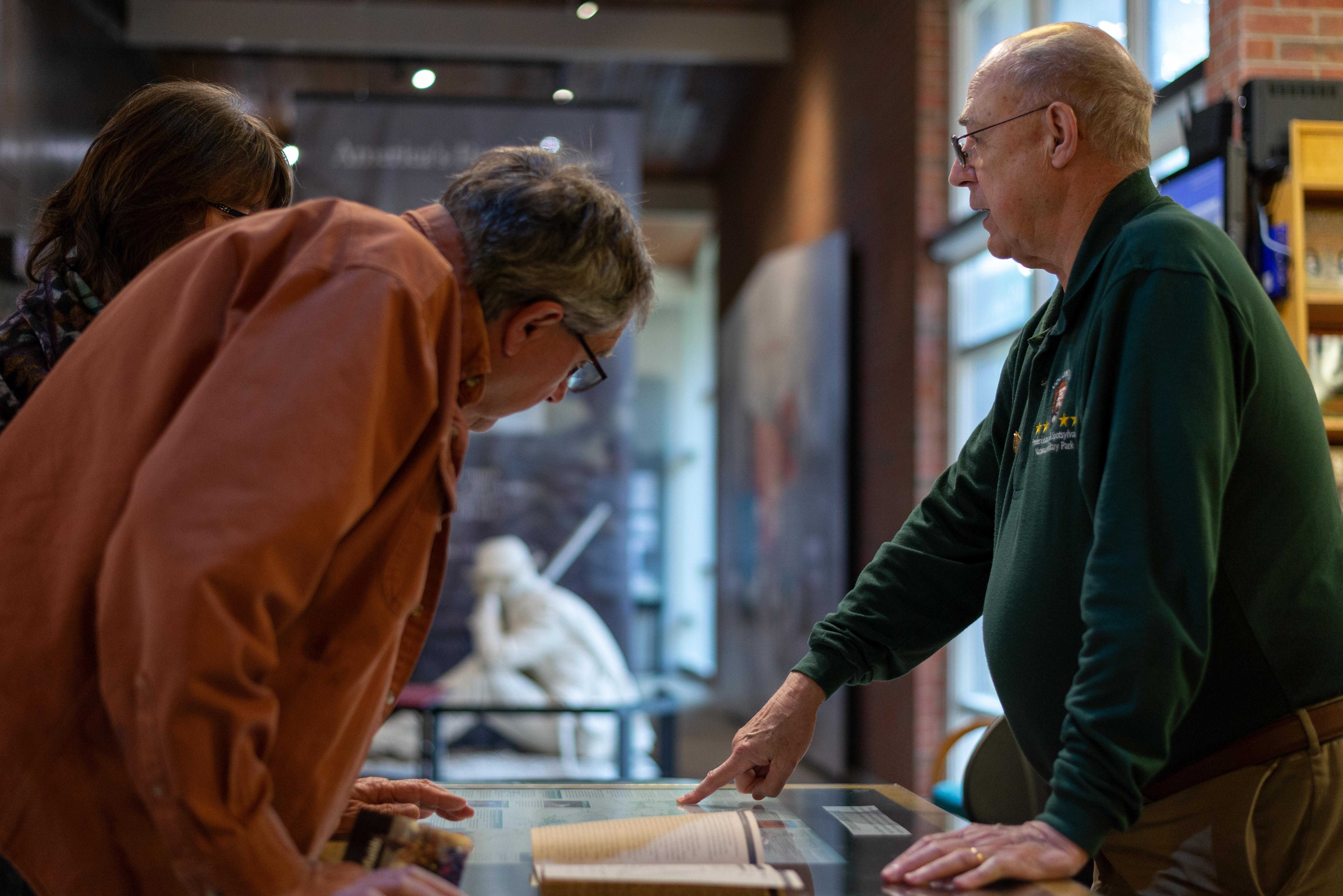 A middle aged couple stand at a visitor center desk and speak with a park service volunteer.