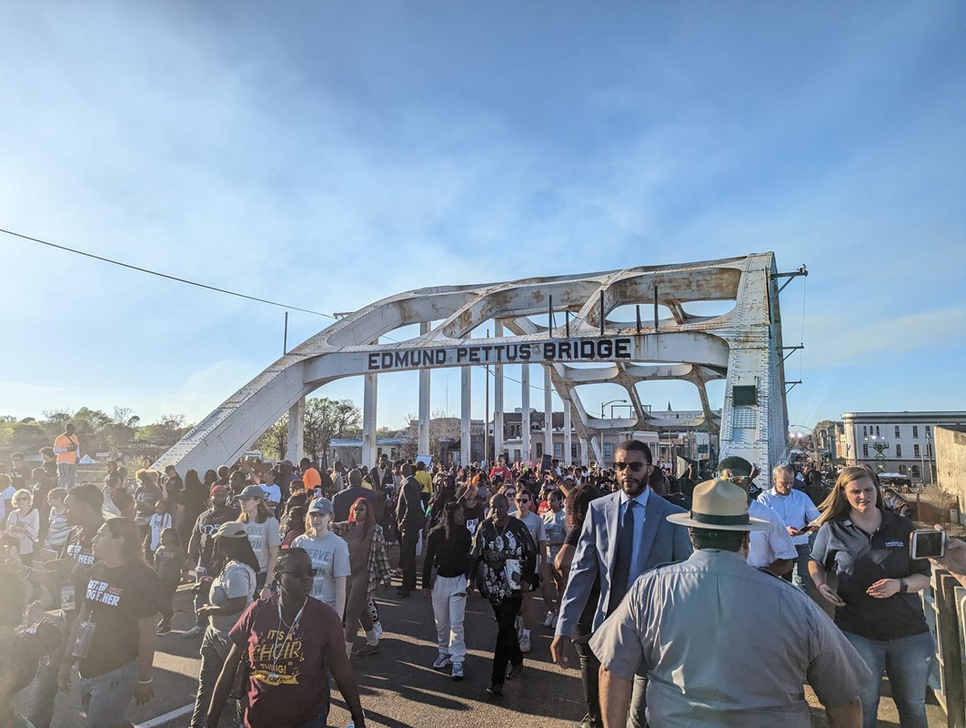 Crowds streaming across a bridge, blue sky and metal beams overhead read, "Edmund Pettus Bridge"