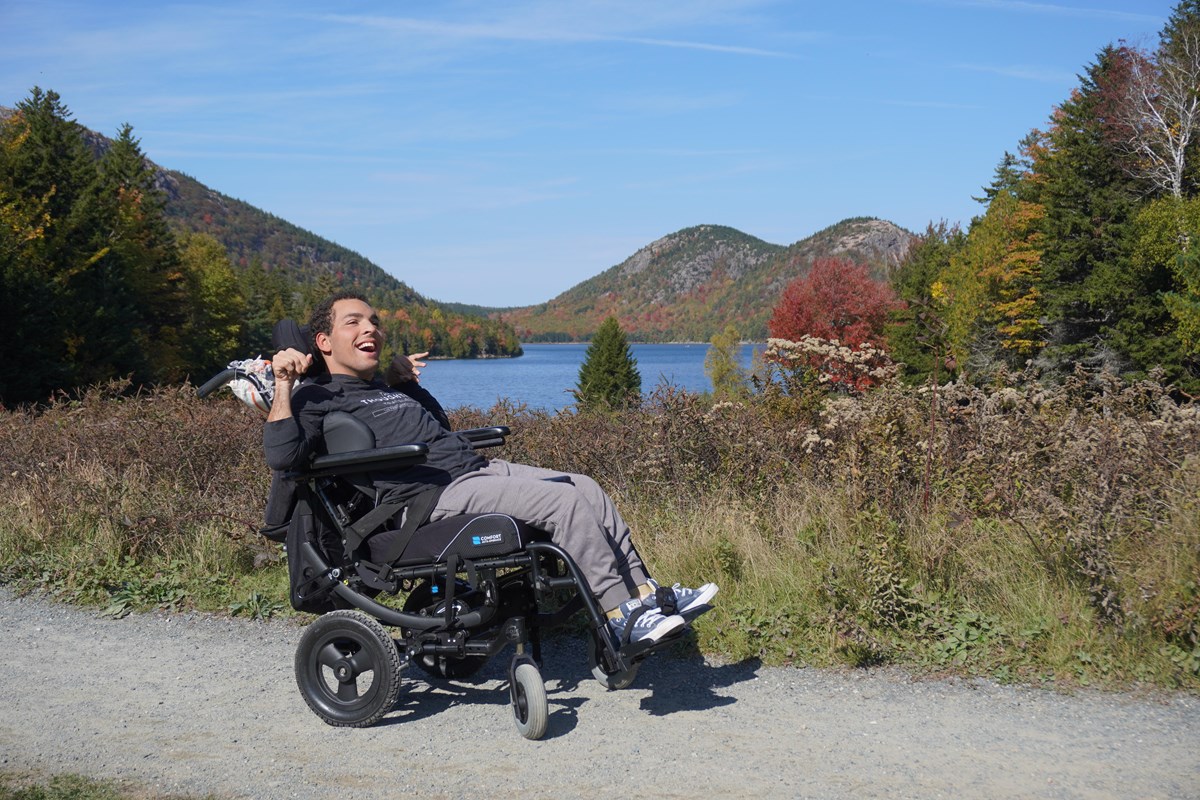 Man in a wheelchair smiles wide while stopped on a path with a pond and mountains in the distance
