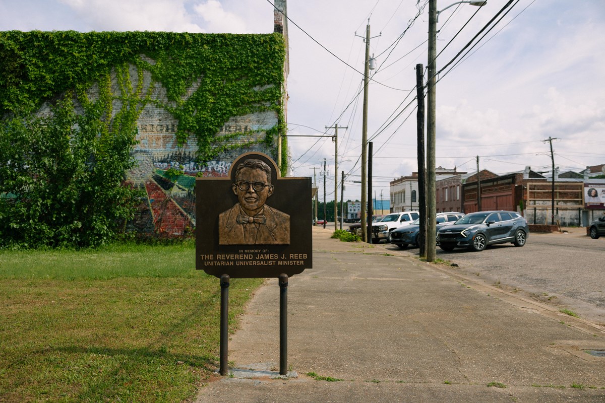A metal plaque depicts a smiling man with a bowtie, a brick building with a mural in the background.