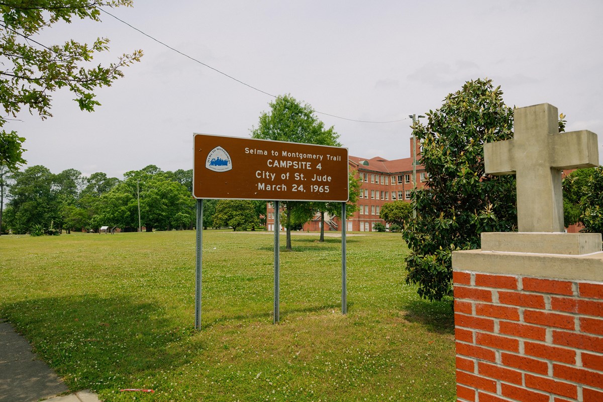 A brown sign reads "Campsite 4. City of St. Jude. March 24, 1965." Brick building in the background.