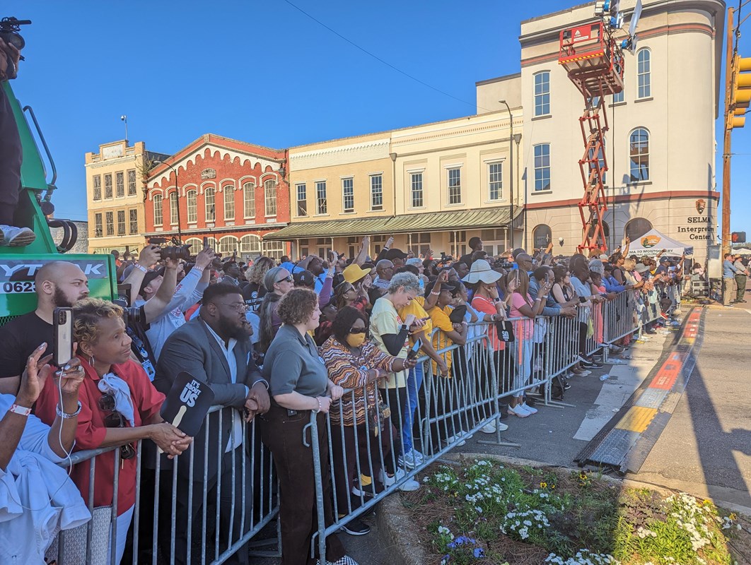 A large crowd of people wait behind a metal barricade on a city street. Buildings in the background.
