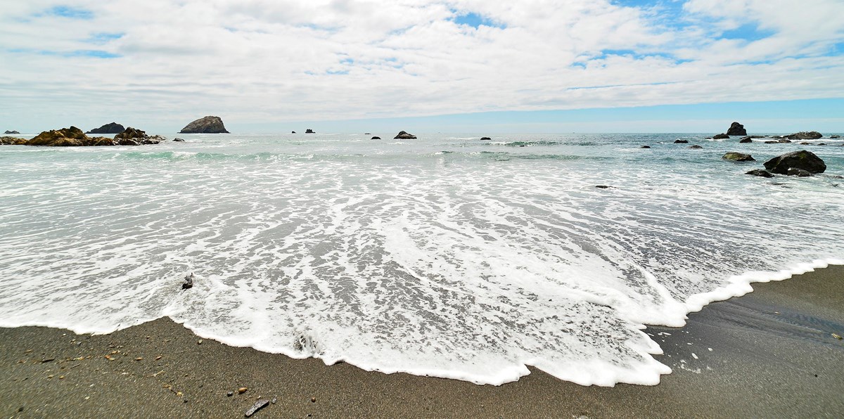 Waves run-up on a beach, and sea stack rocks are seen in the distance.