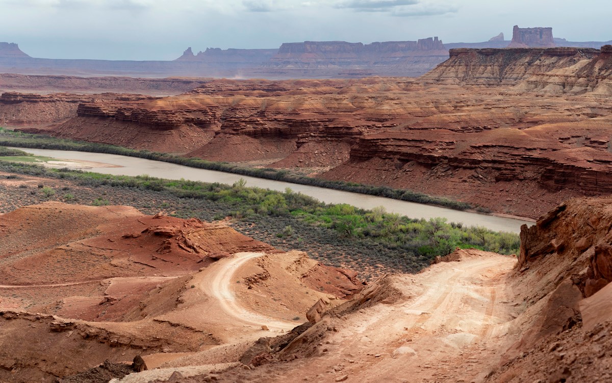 A windy steep dirt road leads up the side of a sandstone cliff. River in background.