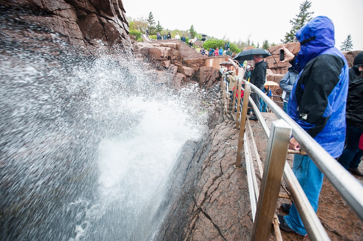 Visitors wearing raincoats and holding umbrellas crowd along a metal railing above a steep rocky inl