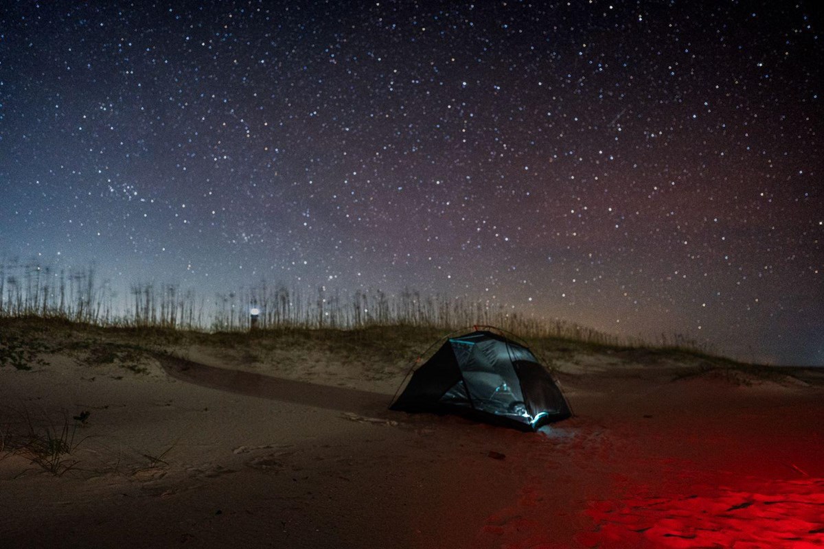 A red light illuminates a tent on the sand, with a star filled sky behind it. 