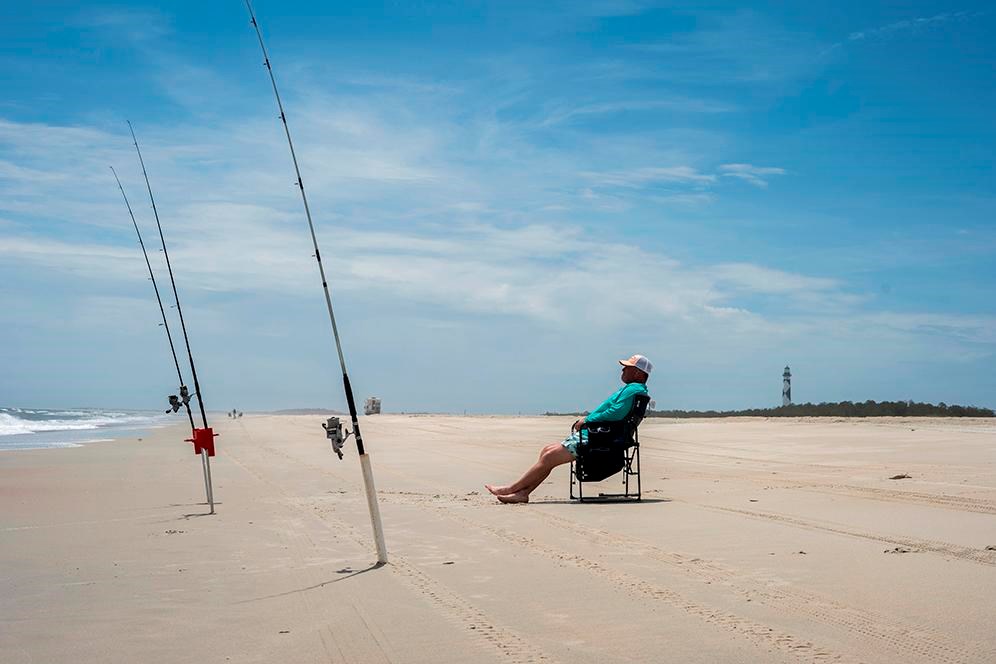 A angler sits in a chair, with two fishing poles in the sand. Lighthouse in background. 