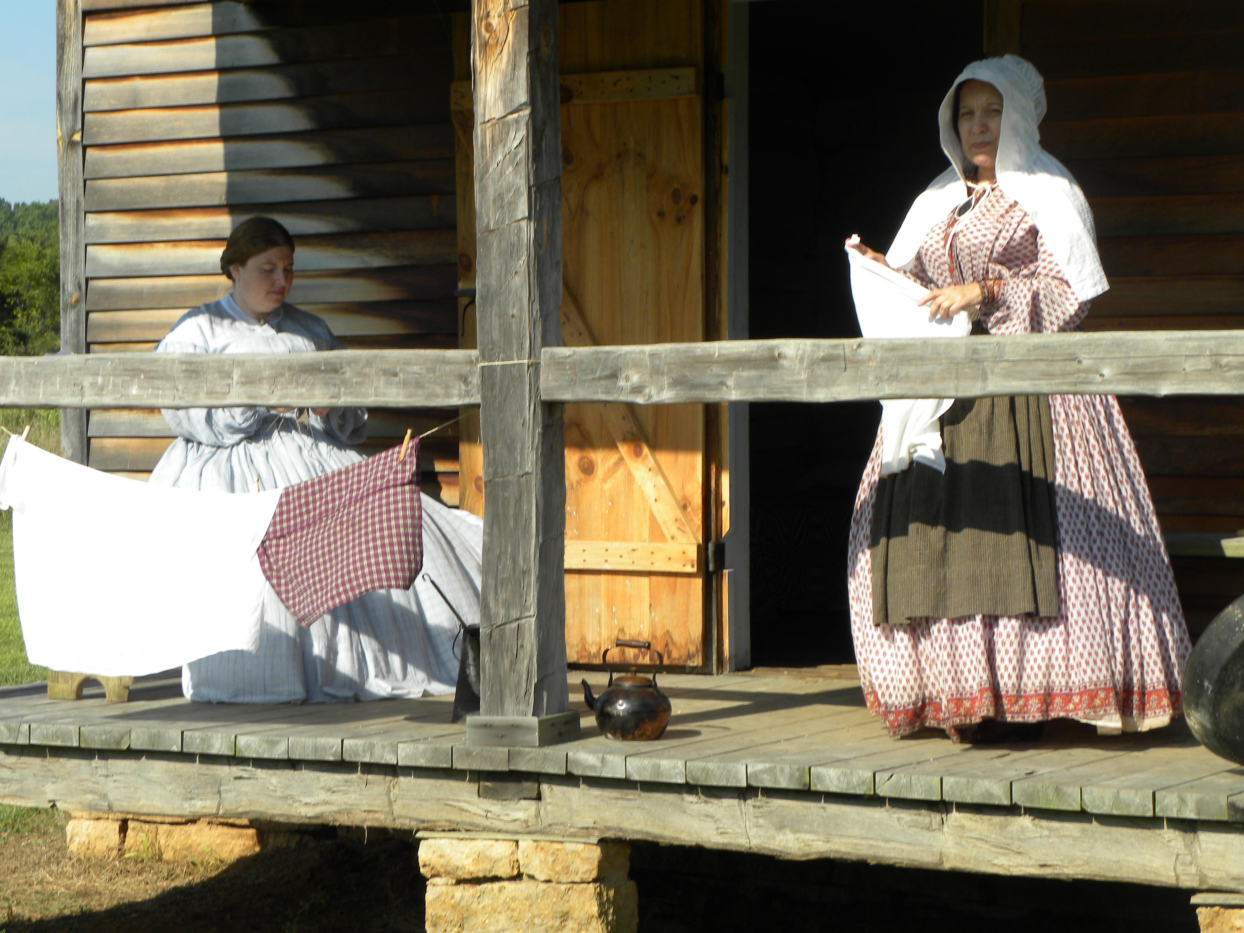 Women in reproduction historic dress sit and stand on porch of old cabin