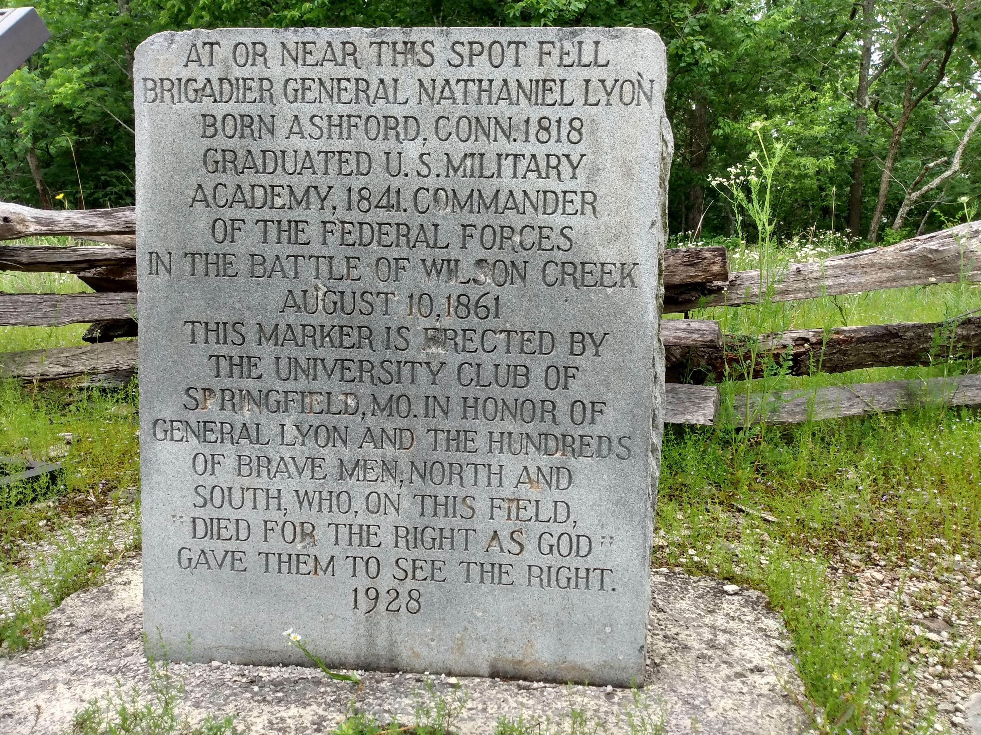 A stone marker with engraving sits in front of a wooden fence and trees