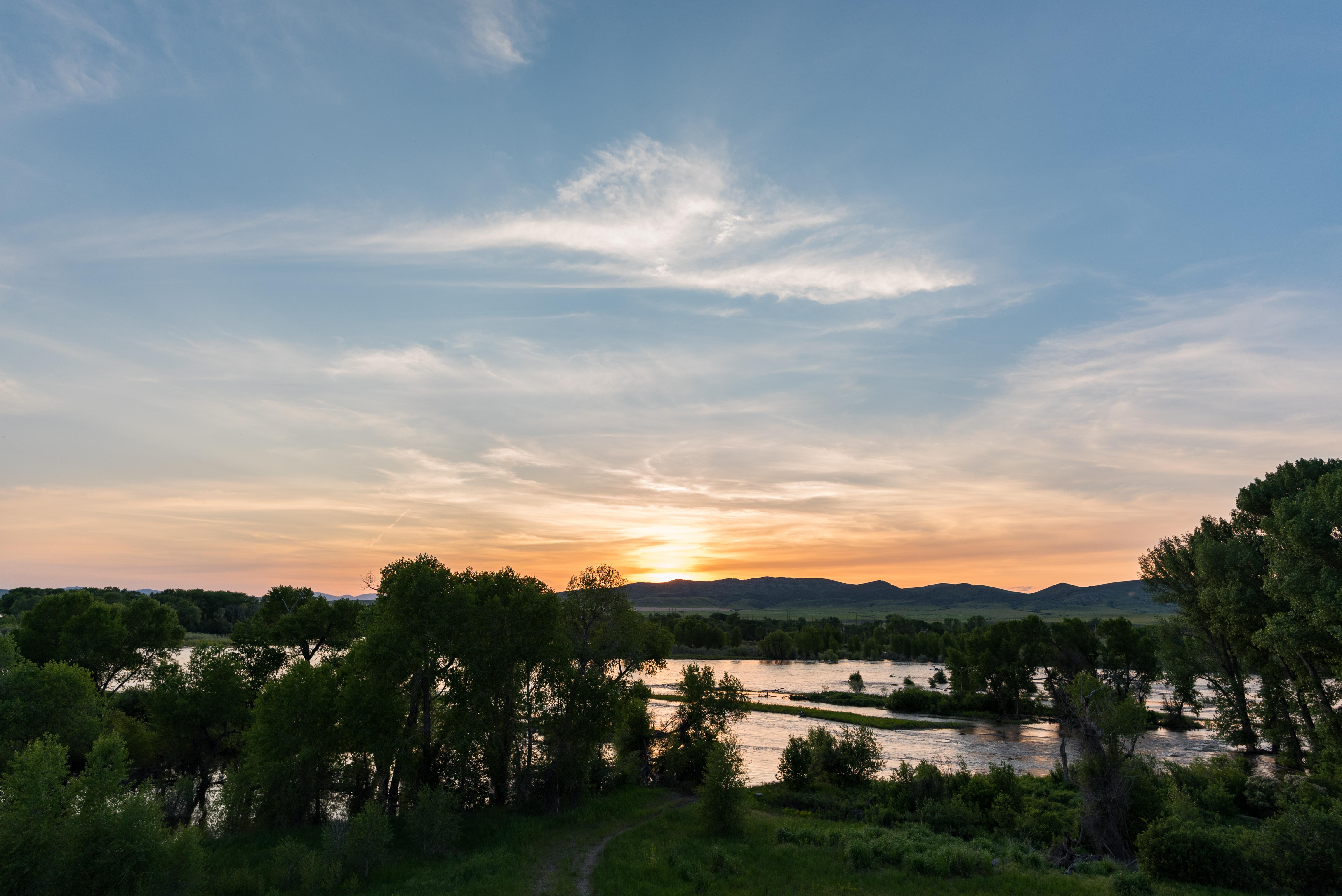 sun dips behind mountains. In the foreground and meandering river catches the light. Leafy trees sur