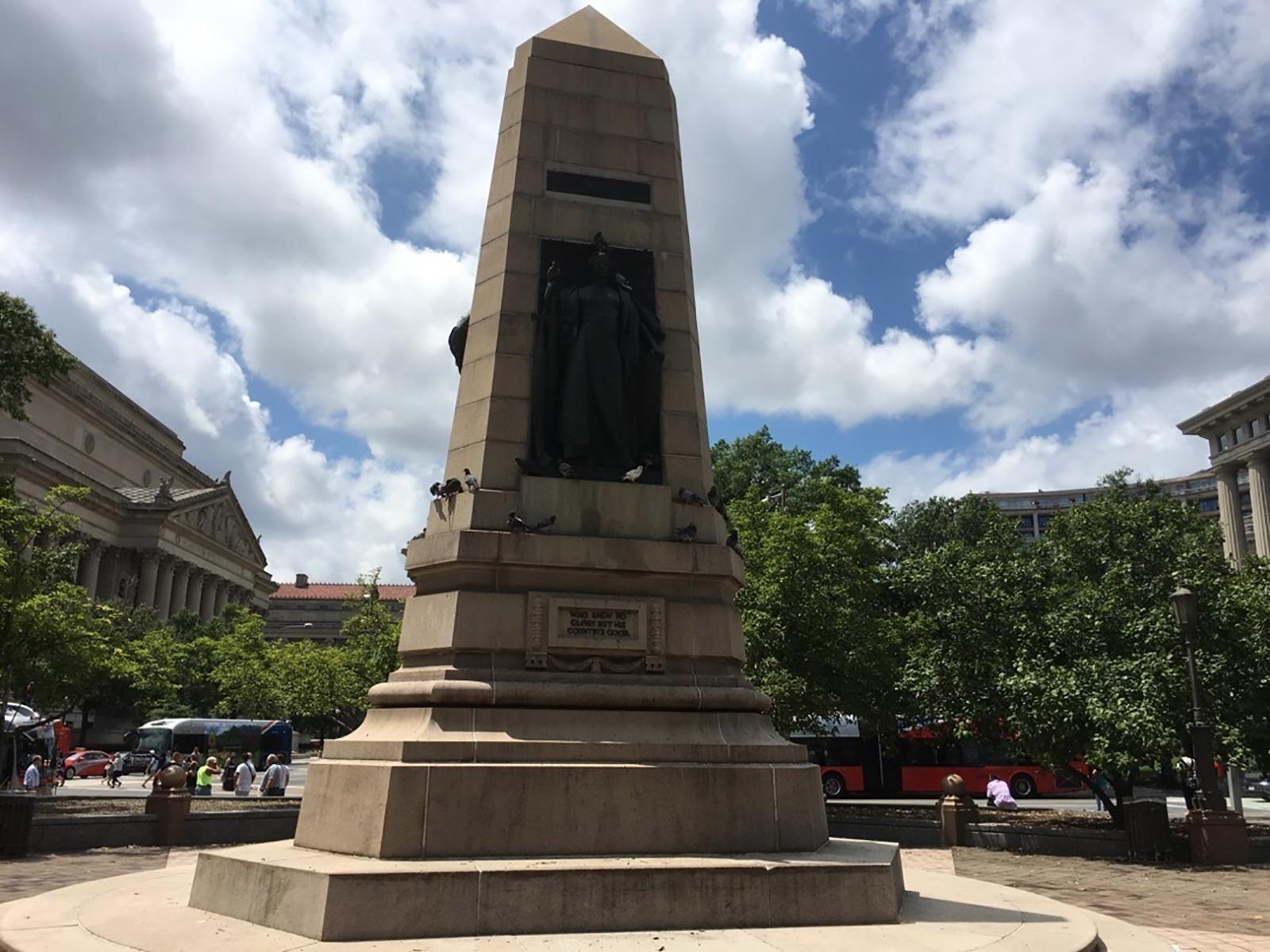 A rather squat, stone obelisk in a plaza with a bronze figure on the face.