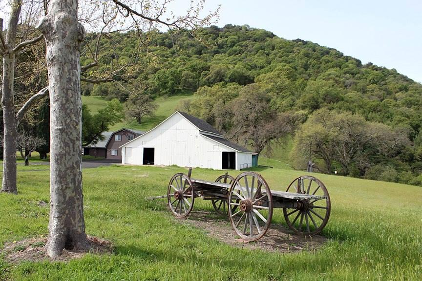 An old barn and an historic wheeled wagon sit in front of a tree-lined hill.