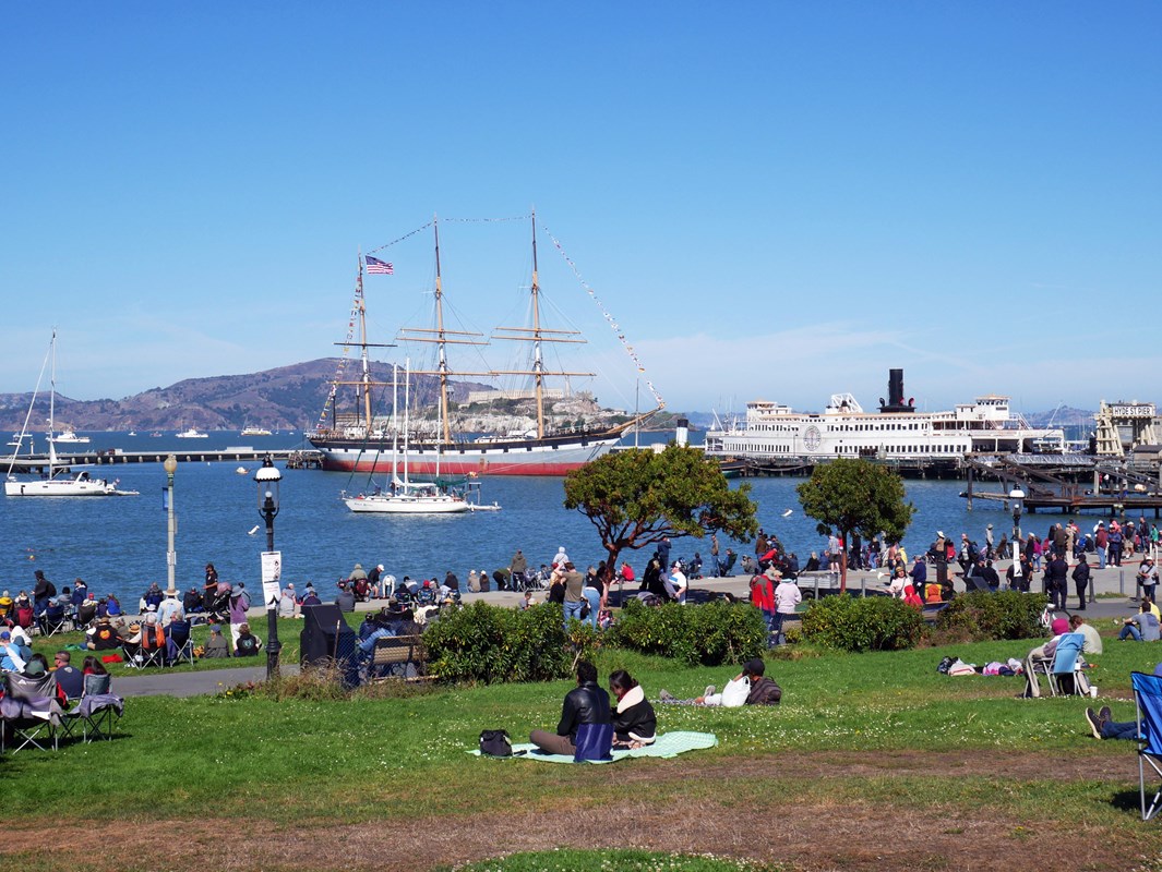 People sitting on a grassy lawn look out over a cove and historic ships in the distance
