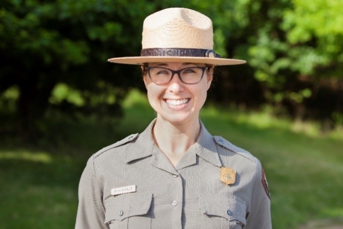 Woman, wearing black-framed eyeglasses and a broad-brimmed hat, stands smiling in front of brilliant green foliage.