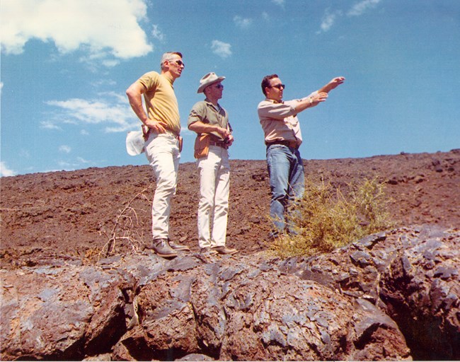 two men standing on dark blueish lava rocks and looking into the distance to where a third man is pointing