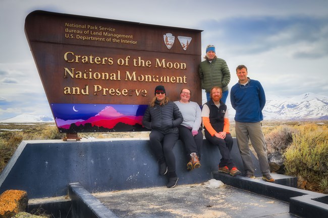 Group of people sit and stand in front of the entrance sign for Craters of the Moon National Monument