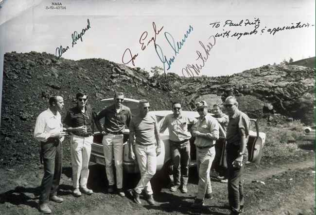 Eight men stand in front of a car parked amongst the lava flows. Image also contains four signatures with an acknowledgement.