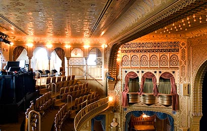 Interior view of an elaborately painted theater, looking across balcony seats toward box seats and the stage on the right.