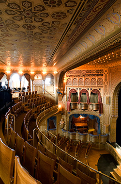 Interior of an elaborately painted theater, looking across balcony seats toward box seats and the stage on the right.