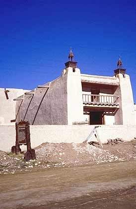 This is an image of an adobe building that has twin towers, a parapet, and various blocks of different sizes and heights that make up its overall massing. It has a recessed balcony and two doorways in the visible central block. The roofing employs vigas. The image rolls over to provide the correct answer. Screen Reader users see text-only link, left, for answer. Photo: NPS files.