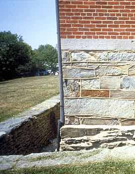 This is a close-up image of the corner of a masonry building. The foundation is formed of large pieces of broken  stone with raised mortar joints. The wall above is made up of roughly rectangular stones that vary in size, color, and texture.  Above that is a narrow band of cut and dressed stones with thin mortar joints. Finally, the main building walls are composed of bricks, uniform in color,  with airly generous mortar joints. Photo: NPS files.