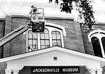 This is an image of a worker on a cherry picker painting the historic wood cornice. A sign on the front of the building says 'Jacksonville Museum.' Photo: Courtesy, Brad Linder, SOHS.