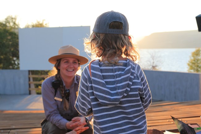 A child with blonde curly hair stands facing a park ranger. Amphitheater seats, a screen, and body of water are behind them.