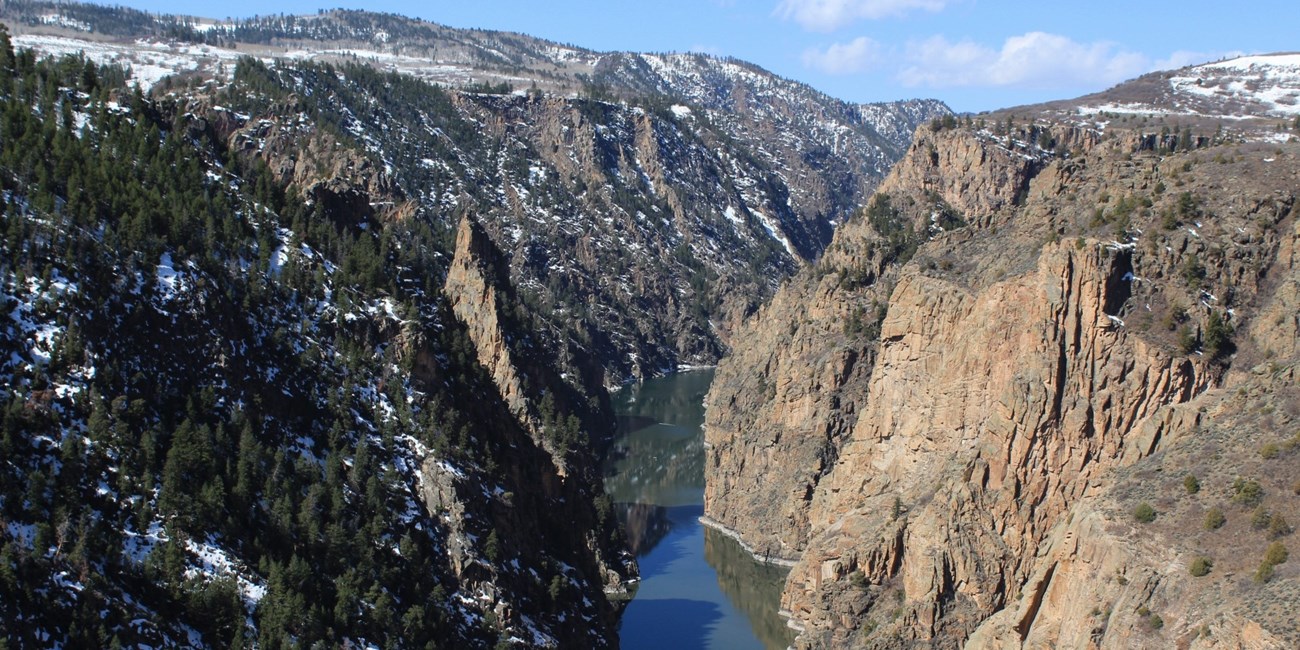 View from above looking into a narrow reservoir between steep canyon walls. Light snow covers the canyon rim and above.