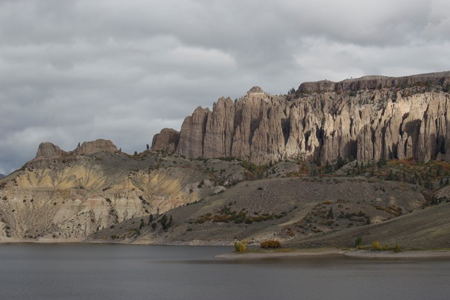 Greyish brown pinnacle spires on the side of a mesa. Water is below the pinnacles.