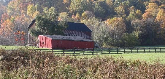 Two-tiered red barn structures sit in front of a wooded hillside in fall colors; a wooden fence along a green field in the foreground.