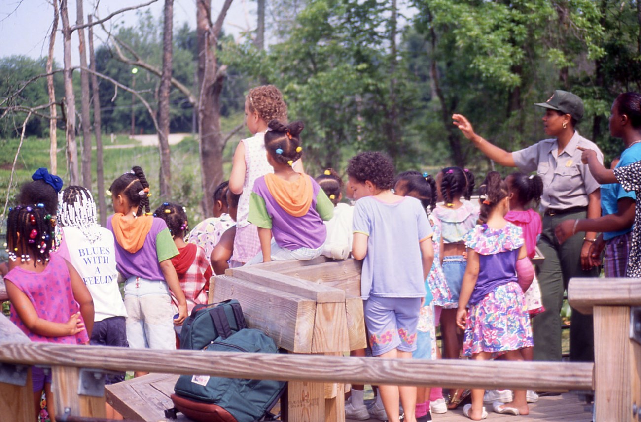 An African American woman in uniform speaks while a group of at least 18 Black girls look over a railing into a wetland.