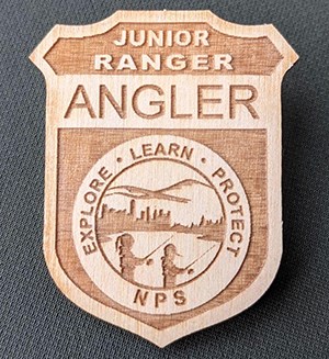 A light brown wooden badge on gray background. Badge reads "Junior Ranger Angler, Explore, Learn, Protect, NPS" and has an illustration of two kids fishing.