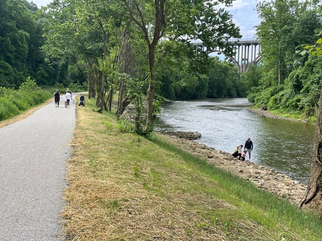People walk pets down a wide, flat trail on a bank next to a river; a tall arched bridge in the distance.